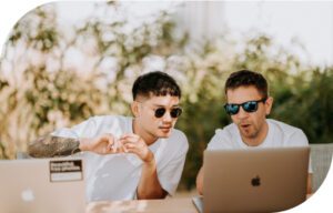 Dos hombres con gafas de sol sentados en una mesa con dos computadoras